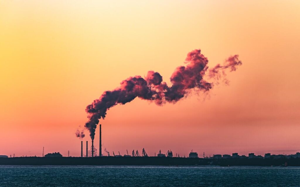 Body of water under cloudy sky during sunset with air pollution venting from smoke stack.